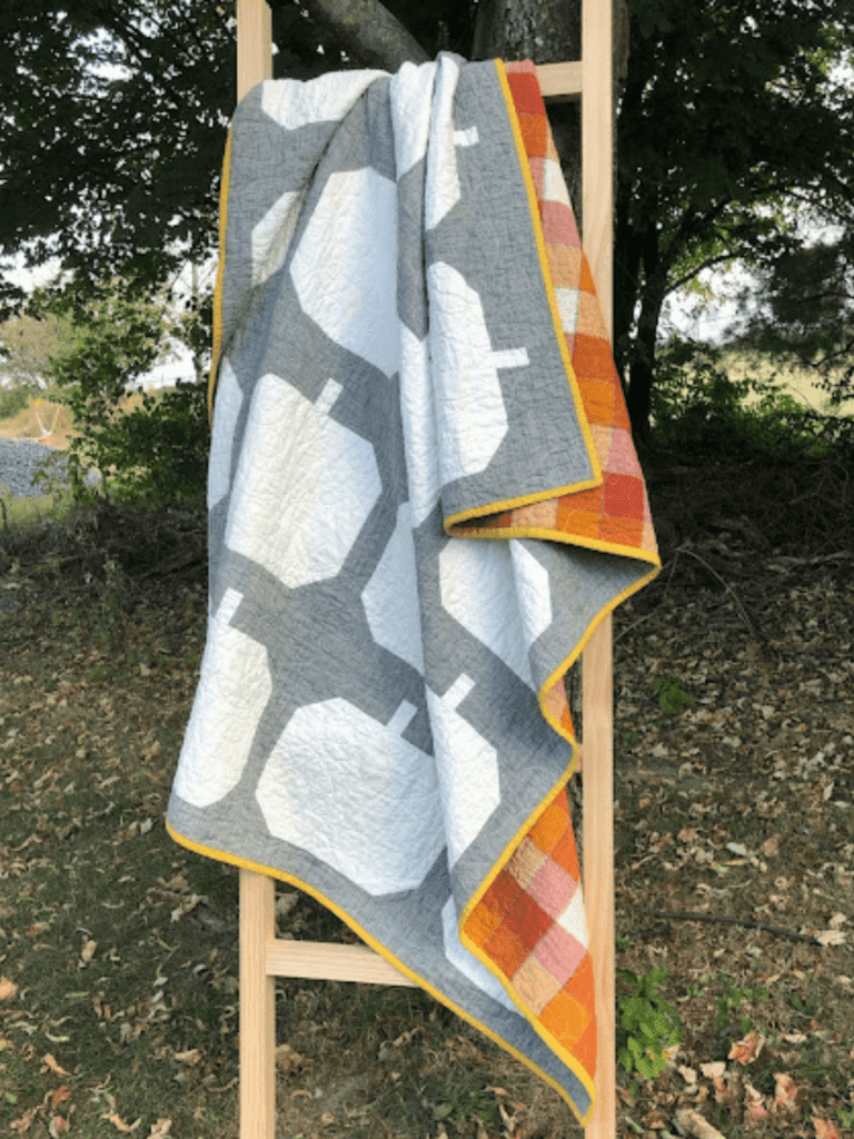 Grey quilt with white pumpkins and an orange plaid backing hanging on a ladder outside.